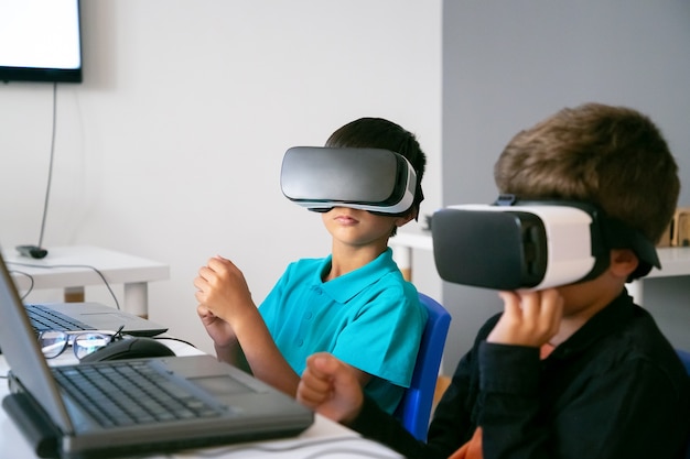 Petits garçons utilisant un casque VR et assis à table avec ordinateur portable