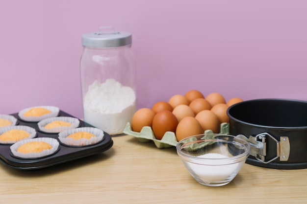 Petits gâteaux cuits au four dans une plaque à pâtisserie avec des ingrédients sur un bureau en bois sur fond rose