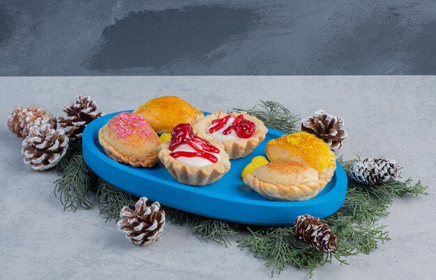 Petits gâteaux, petits pains et bonbons à la gelée sur un plateau bleu décoré de feuilles de pin et de cônes sur une surface en marbre