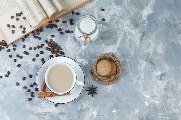 Un peu de café avec des biscuits, des grains de café, un livre, du lait, des épices dans une tasse sur fond de plâtre gris, vue de dessus.