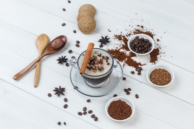 Un peu de café avec des grains de café, du café moulu, des épices, des biscuits, des cuillères en bois dans une tasse sur fond en bois, vue grand angle.