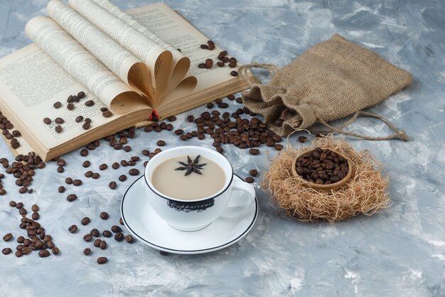 Un peu de café avec des grains de café, livre, sac dans une tasse sur fond de plâtre gris, vue grand angle.