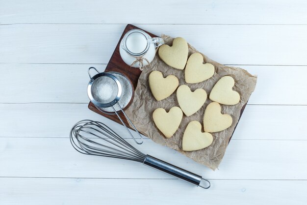 Un peu de lait avec des biscuits, du sucre en poudre, une passoire, un fouet dans une cruche sur fond blanc et planche à découper, vue de dessus.