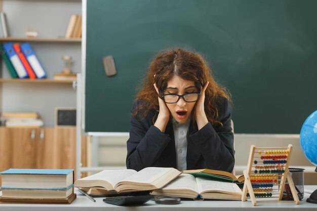 peur attrapé la tête jeune enseignante portant des lunettes lisant un livre assis au bureau avec des outils scolaires en classe