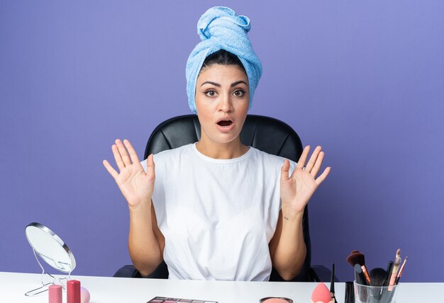 Peur belle femme assise à table avec des outils de maquillage cheveux enveloppés dans une serviette montrant un geste d'arrêt