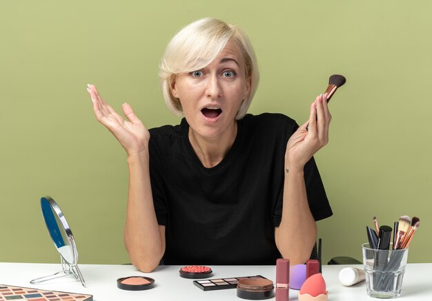 Peur belle jeune fille assise à table avec des outils de maquillage tenant un pinceau à poudre isolé sur fond vert olive