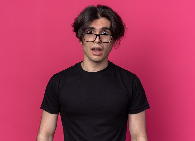 Peur de cheveux sauvages jeune beau mec portant un t-shirt noir et des lunettes isolés sur un mur rose