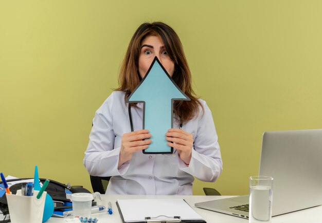 Peur de femme médecin d'âge moyen portant une robe médicale avec stéthoscope assis au bureau de travail sur un ordinateur portable avec des outils nedical visage couvert avec marque de direction avec espace de copie