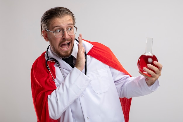 Peur de gars jeune super-héros portant une robe médicale avec stéthoscope et lunettes tenant une bouteille en verre de chimie remplie de liquide rouge isolé sur fond blanc