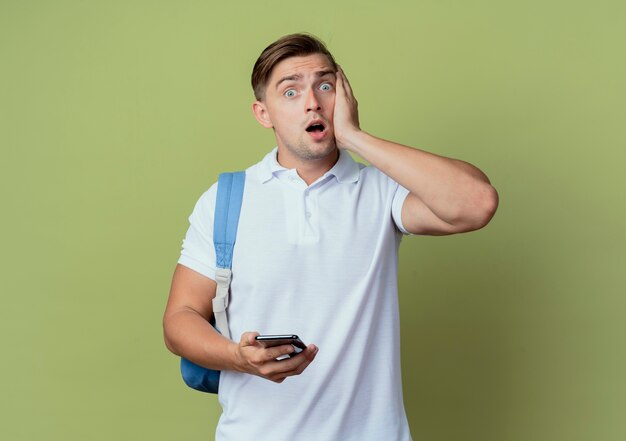 Peur jeune beau étudiant masculin portant un sac à dos tenant le téléphone et mettant la main sur la joue isolé sur vert olive