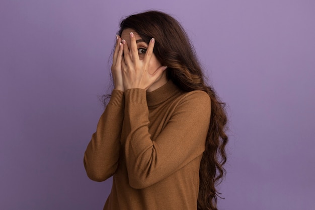 Peur de jeune belle fille portant un pull à col roulé marron visage couvert avec les mains isolées sur le mur violet