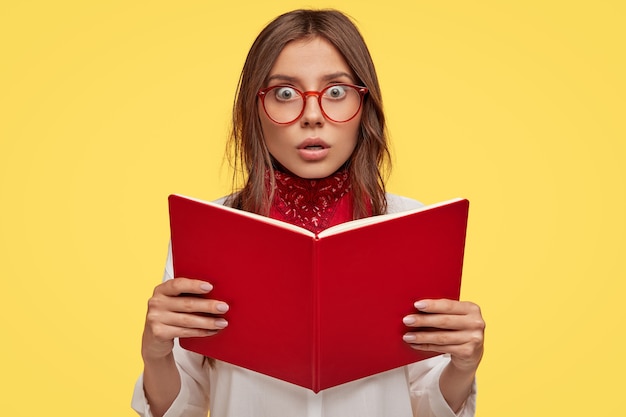 Peur de jeune brune avec des lunettes posant contre le mur jaune