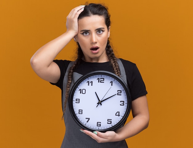 Peur de jeune femme coiffeur en uniforme tenant horloge murale et mettre la main sur la tête isolé sur mur orange