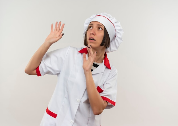 Peur de jeune femme cuisinier en uniforme de chef à la recherche de côté avec les mains levées isolé sur mur blanc