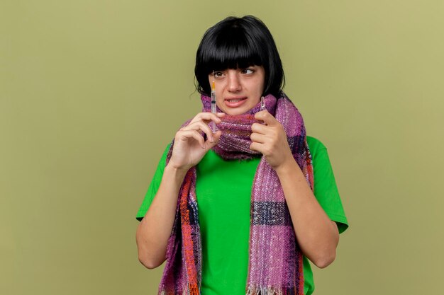Peur de jeune femme malade portant un foulard tenant la seringue et l'ampoule en regardant la seringue isolée sur le mur vert olive avec espace copie