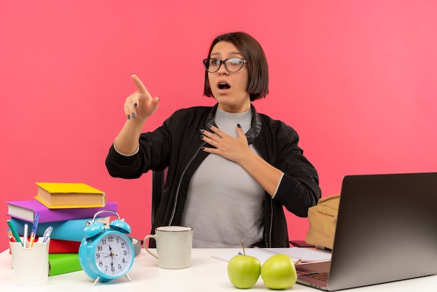 Peur De Jeune Fille étudiante Portant Des Lunettes Assis Au Bureau à Faire Ses Devoirs à La Recherche Et Pointant Sur Le Côté Avec La Main Sur La Poitrine Isolée Sur Le Mur Rose