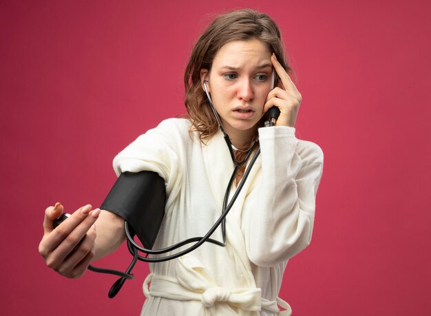 Peur de jeune fille malade à côté portant une robe blanche mesurant sa propre pression avec sphygmomanomètre mettant la main sur le temple isolé sur rose