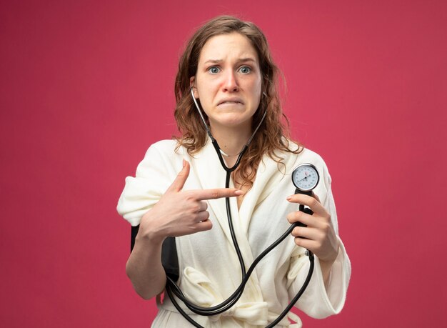 Peur de jeune fille malade portant une robe blanche mesurant sa propre pression avec sphygmomanomètre isolé sur rose