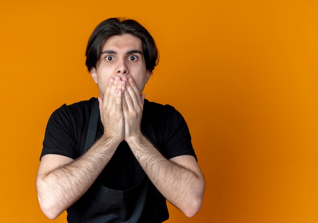 Peur de jeune homme beau coiffeur en uniforme couvert de bouche avec les mains isolés sur orange avec copie espace