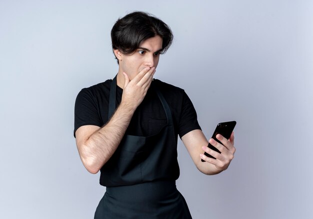 Peur de jeune homme beau coiffeur en uniforme tenant et regardant la bouche couverte de téléphone avec la main sur blanc