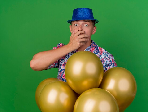 Peur jeune homme de fête portant un chapeau bleu debout derrière des ballons et le visage couvert avec la main isolé sur vert