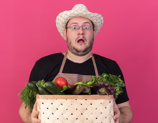 Peur jeune jardinier mâle portant chapeau de jardinage tenant un panier de légumes isolé sur mur rose