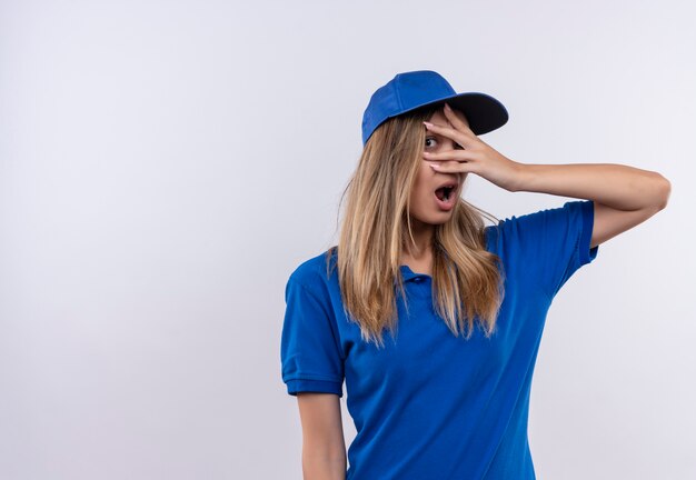 Peur de jeune livreuse portant l'uniforme bleu et les yeux couverts de casquette avec la main isolé sur un mur blanc avec espace de copie