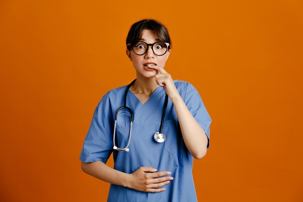 Peur de mettre le doigt sur la bouche jeune femme médecin portant un stéthoscope fith uniforme isolé sur fond orange