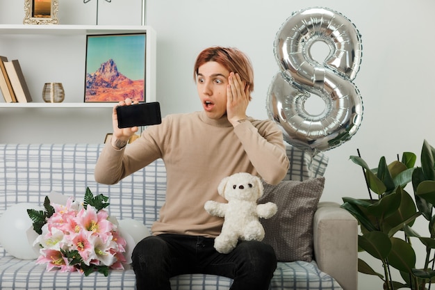 Peur de mettre la main sur la joue beau mec le jour de la femme heureuse tenant un ours en peluche avec un téléphone assis sur un canapé dans le salon