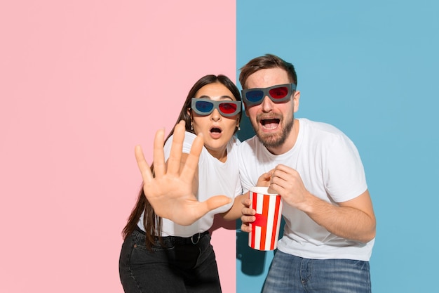 Peur de regarder le cinéma 3D avec du pop-corn. Jeune et heureux homme et femme dans des vêtements décontractés sur un mur bicolore rose, bleu. Concept d'émotions humaines, expression faciale, relations. Beau couple.