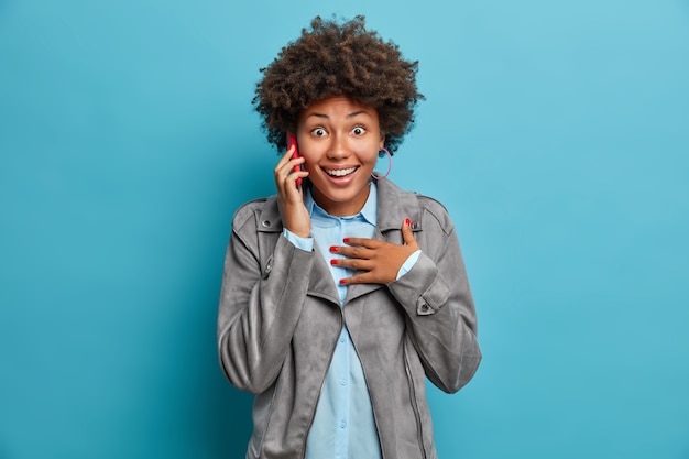 Photo d'une adolescente aux cheveux bouclés heureux surpris d'expression joyeuse parle via téléphone mobile réagit de manière surprenante sur des nouvelles impressionnantes porte une veste grise