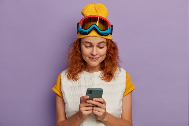 Photo d'une adolescente aux cheveux roux active utilise un téléphone portable pour discuter en ligne, passe des vacances d'hiver dans les montagnes, porte un masque de protection de snowboard, aime voyager et une connexion Internet gratuite