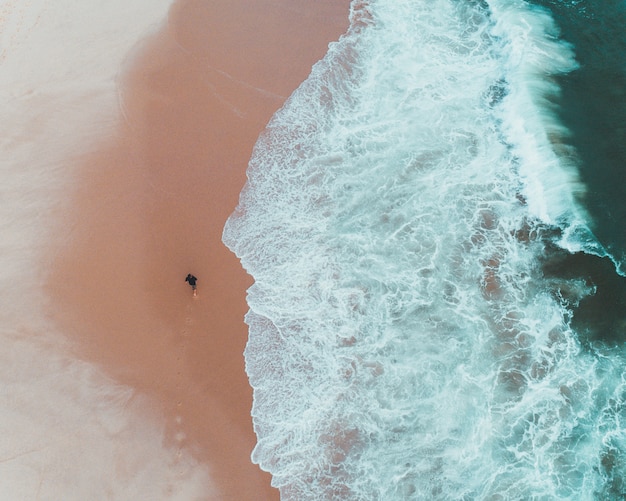 Photo aérienne d'un homme marchant sur la plage aux côtés de belles vagues de l'océan frappant la côte