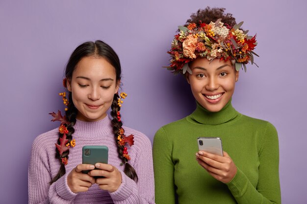 La photo d'amies heureuses tient des smartphones modernes, envoie des messages à des amis, utilise une manière créative de porter des feuilles d'automne, a des expressions joyeuses, porte des pulls.