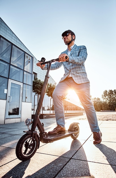 Photo en angle bas d'un homme élégant et intelligent portant des lunettes de soleil et un casque qui conduit son scooter électrique.