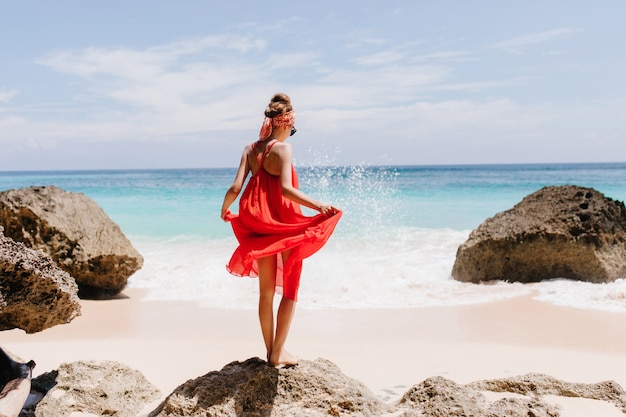 Photo de l'arrière d'une fille bronzée galbée debout sur une grosse pierre. Plan extérieur d'un modèle féminin gracieux jouant avec sa robe rouge et regardant les vagues de l'océan.