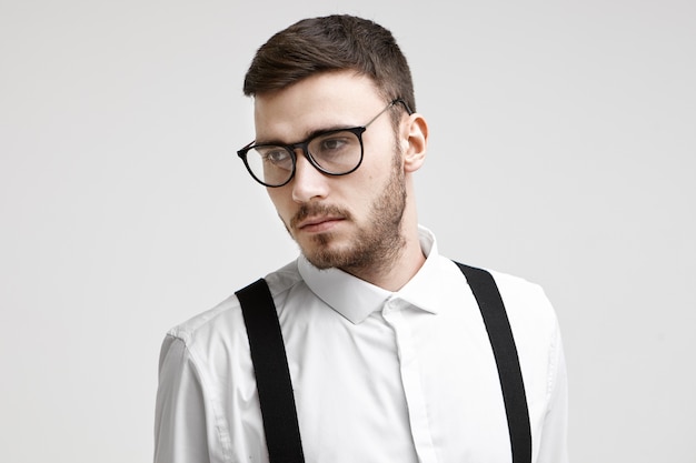 Photo de beau jeune homme d'affaires caucasien avec barbe taillée et moustache debout au mur de bureau blanc, ayant un regard sérieux et réfléchi. Concept d'entreprise, de style et de réussite
