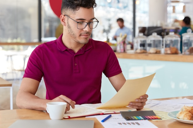 Photo d'un beau jeune homme aux cheveux noirs tient un document, lit des informations, a ouvert un cahier, étudie des graphiques et des diagrammes, porte des vêtements décontractés, boit du café aromatique, pose dans un café confortable