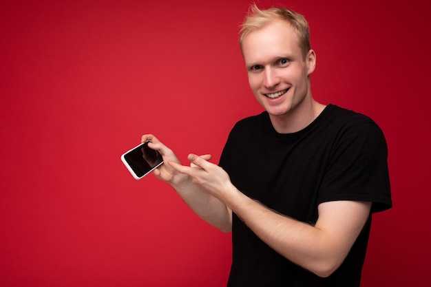 Photo d'un beau jeune homme blond souriant portant un t-shirt noir debout isolé sur rouge