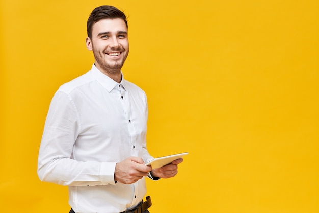 Photo de beau jeune homme confiant en chemise blanche tenant une tablette numérique générique et souriant largement, appréciant jouer à des jeux à l'aide d'une application en ligne. Technologie, divertissement et jeux