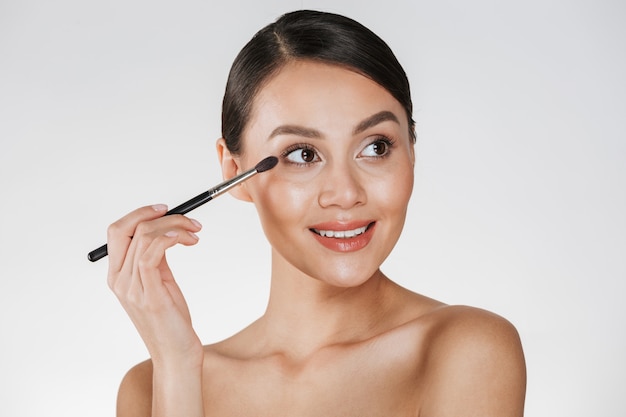 Photo de beauté de l'adorable femme brune en détournant les yeux et souriant tout en appliquant des cosmétiques avec un pinceau pour le fard à paupières, isolé sur blanc