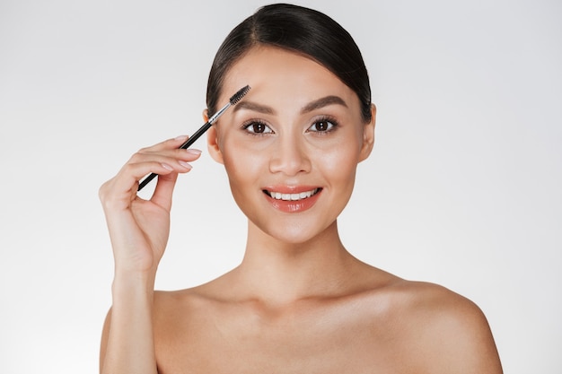 Photo de beauté de jolie jeune femme aux cheveux en chignon à la recherche à la caméra et peigner ses sourcils avec une brosse, isolé sur blanc