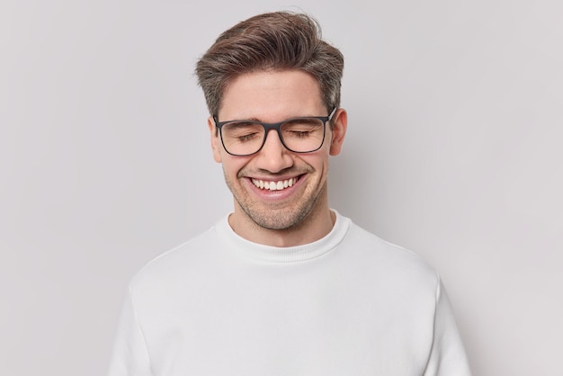 Photo d'un bel homme gai aux cheveux noirs sourit avec joie les yeux fermés exprime des émotions positives rêverie à propos de quelque chose porte des lunettes et un pull décontracté isolé sur un mur blanc