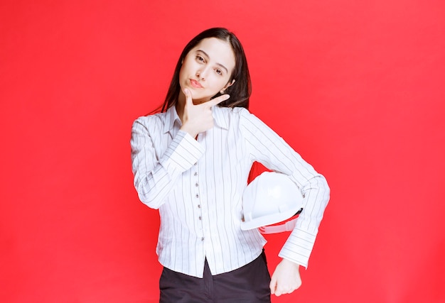 Photo d'une belle femme d'affaires tenant un chapeau de sécurité sur un mur rouge.