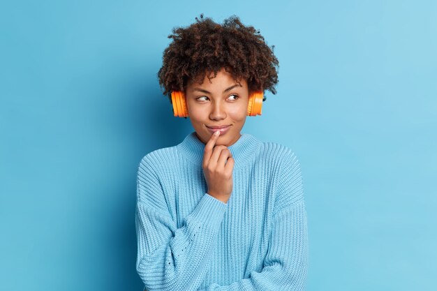 Photo de belle femme afro-américaine se tient pensivement à l'intérieur écoute une chanson agréable qui lui apporte de bons souvenirs du passé porte des écouteurs occasionnels sur les oreilles