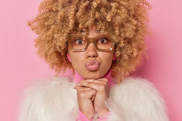 La photo d'une belle femme garde les lèvres pliées a une expression romantique des cheveux blonds bouclés vêtus d'un manteau de fourrure blanc isolé sur fond rose attend le baiser. Modèle féminin affectueux à l'intérieur