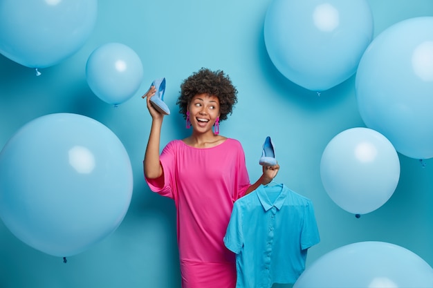 La photo d'une belle femme joyeuse choisit quoi porter, sélectionne une tenue bleue pour un événement spécial, tient des chaussures à talons hauts et une chemise sur un cintre, pose contre un mur décoré. Collection de vêtements