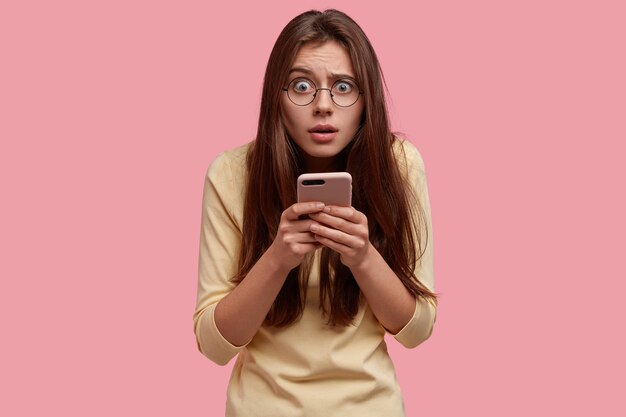 Photo d'une belle femme perplexe a une expression faciale indignée, porte un téléphone intelligent dans les mains, tape des messages, habillé avec désinvolture