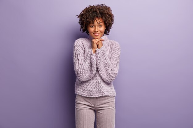 Photo d'une belle femme ravie garde les mains sous le menton, sourit joyeusement, écoute attentivement des informations agréables, porte un pull violet d'hiver, pose à l'intérieur. Concept d'expressions faciales.