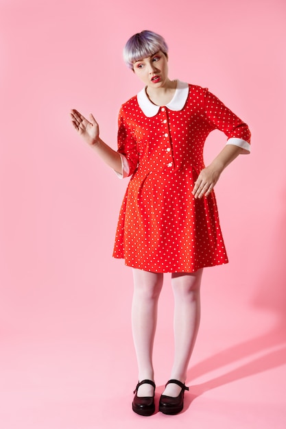 Photo de belle fille de poupée avec des cheveux courts violet clair portant une robe rouge sur un mur rose
